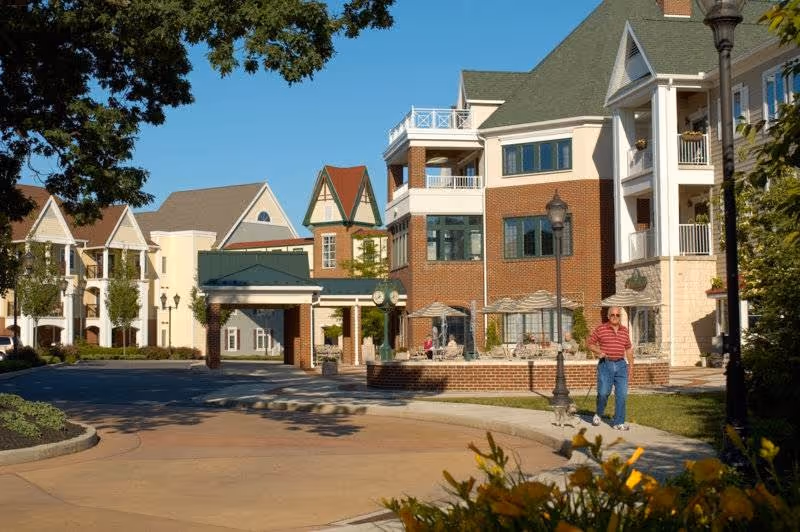 Exterior view of a senior living community with multi-story buildings featuring brick and light-colored siding. There are outdoor seating areas with umbrellas, a covered entrance, and a man walking on the sidewalk. Trees and landscaping are visible under a clear blue sky.