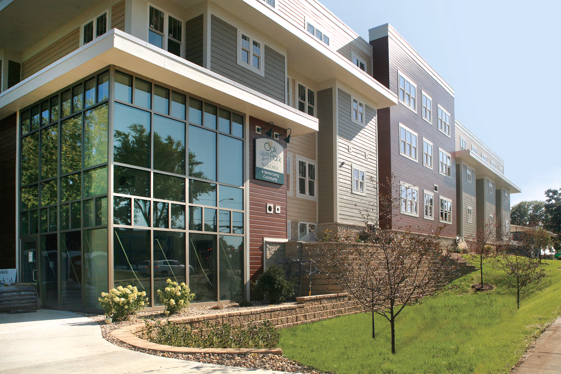 Exterior view of Oak Park Place Nakoma, a senior living community building with large glass windows, beige and brown siding, and a landscaped area with small trees and shrubs in front.