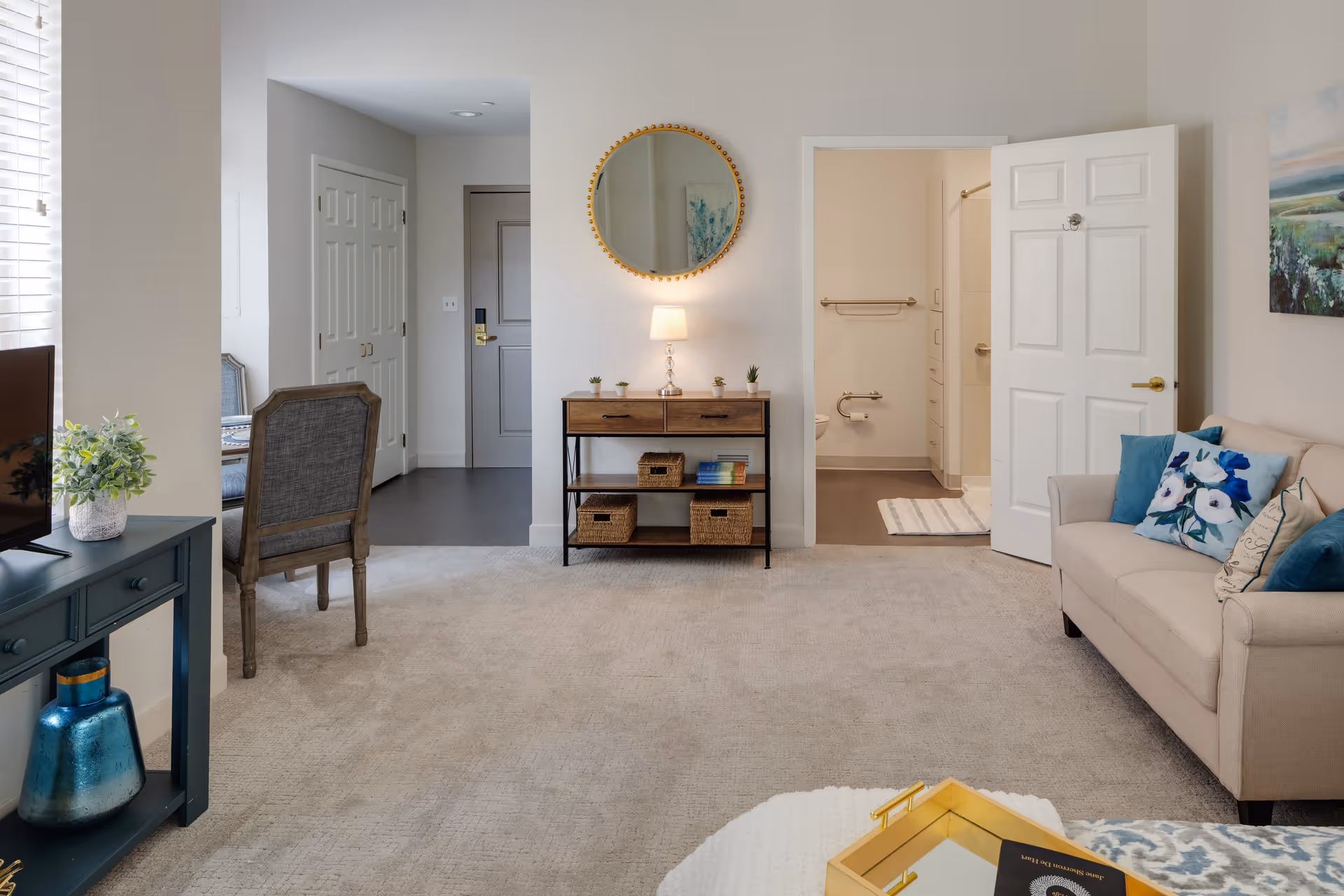 A cozy living room area with a beige sofa adorned with blue and floral pillows on the right, a small wooden console table with baskets and a lamp against the wall, and a round mirror hanging above it. To the left, there is a dark green TV stand with a plant and decorative items. The room has beige carpeting and light-colored walls. An open door reveals a bathroom with grab bars and a towel rack. A dining area with chairs is partially visible through an open doorway on the left.