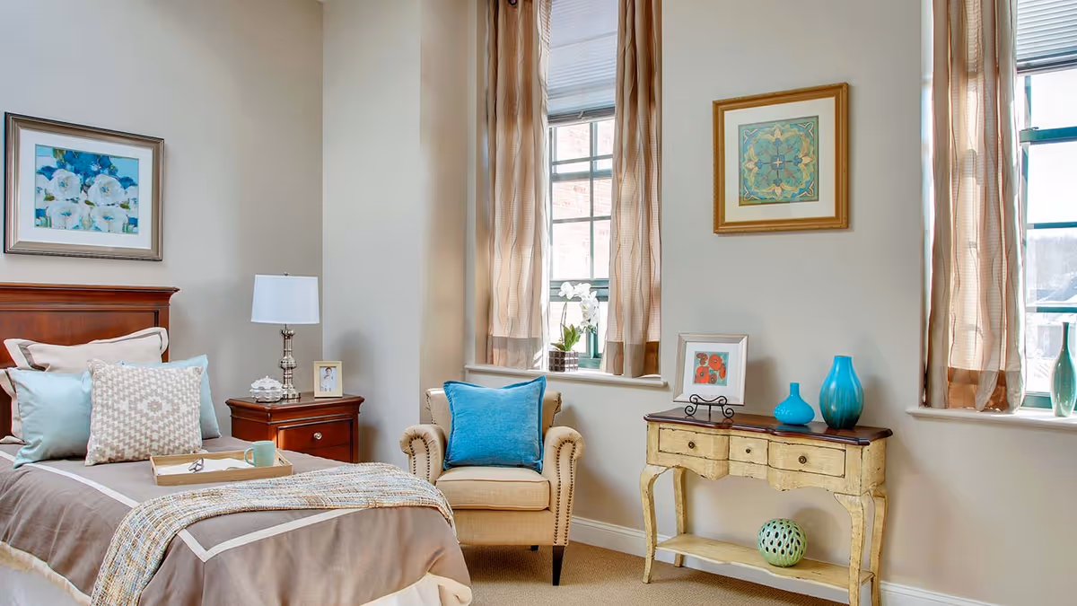 A cozy bedroom featuring a single bed with multiple pillows and a beige and brown comforter. Next to the bed is a wooden nightstand with a lamp, a small framed photo, and decorative items. There is a beige armchair with a blue pillow near two windows with light brown curtains. A vintage-style wooden console table with decorative vases and framed art is positioned under the windows. The walls are light-colored and adorned with framed artwork.