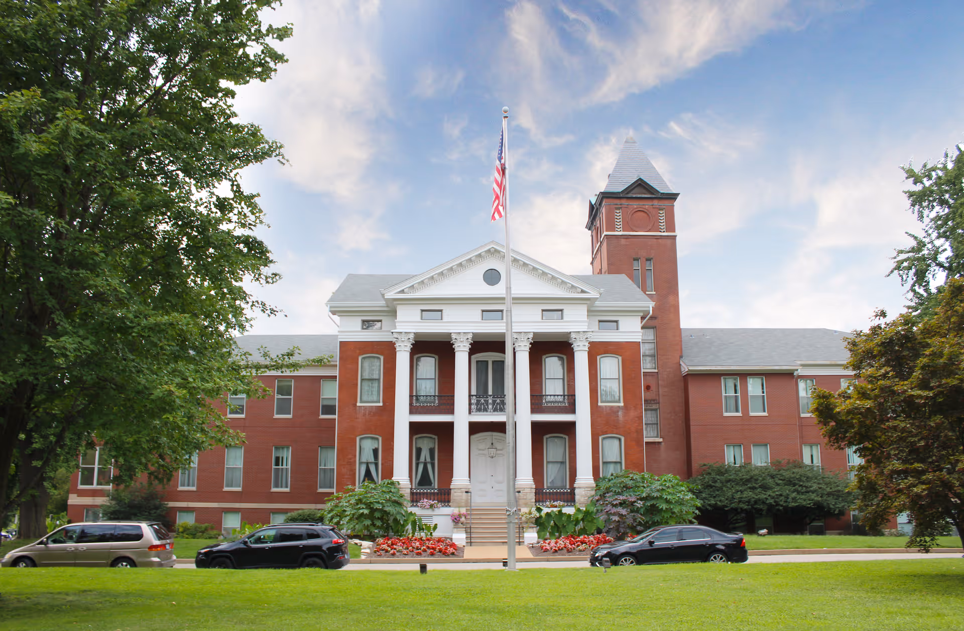 Front exterior view of a large red brick building with white columns and a tall tower, surrounded by green trees and lawn, with an American flag on a flagpole in front and several parked cars.