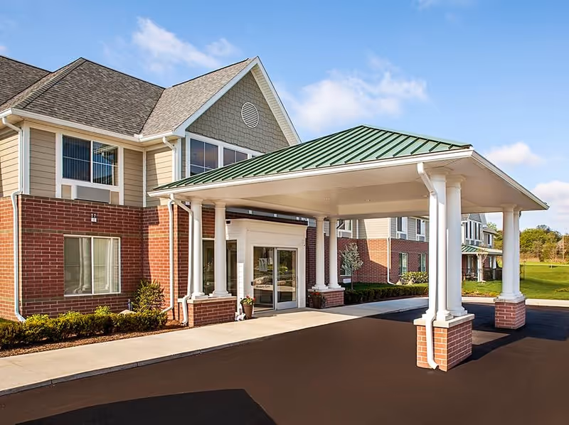 Exterior view of Belmar Oakland senior living facility entrance with a covered drop-off area supported by white columns and a green metal roof, brick and beige siding on the building, and a clear blue sky.
