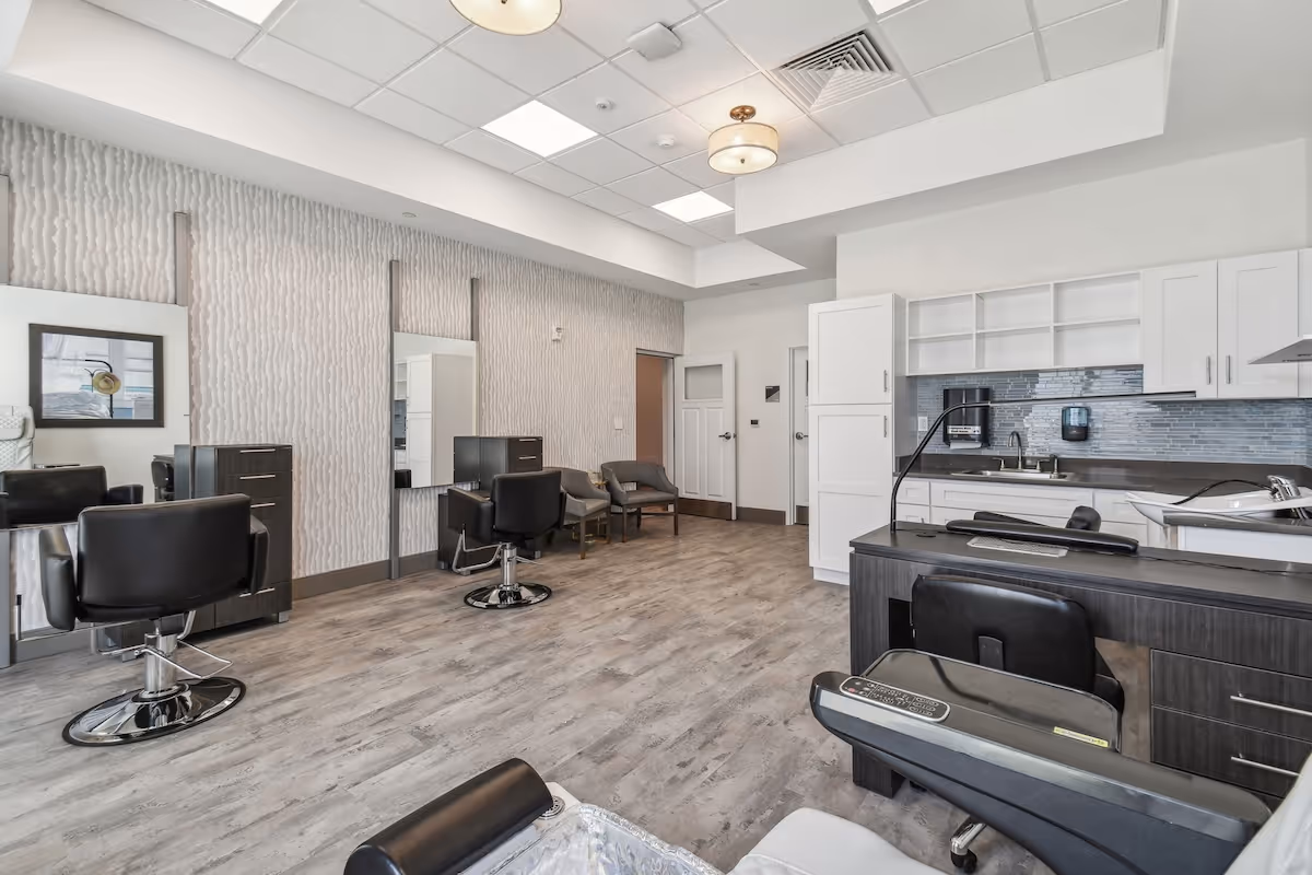 Interior view of a modern salon area with black styling chairs, mirrors on the wall, a manicure station, and a kitchenette with white cabinets and a gray backsplash. The room has light wood flooring and a textured accent wall.