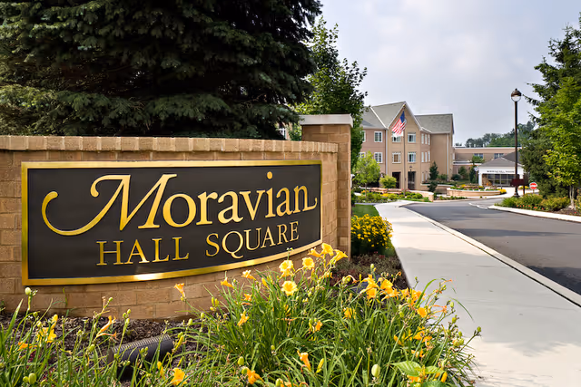 Entrance sign for Moravian Hall Square with yellow flowers in the foreground, a sidewalk, and a multi-story building with an American flag in the background under a partly cloudy sky.