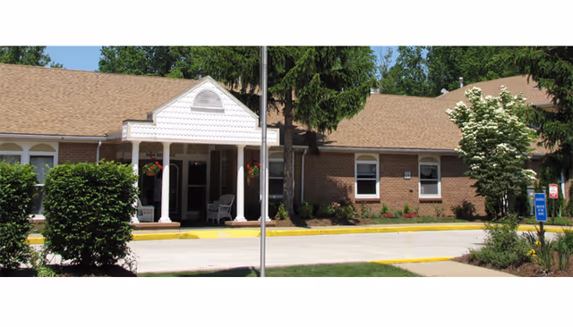 Exterior view of a single-story brick building with a covered entrance supported by white columns. There are bushes, trees, and a small garden area in front of the building under a clear blue sky.