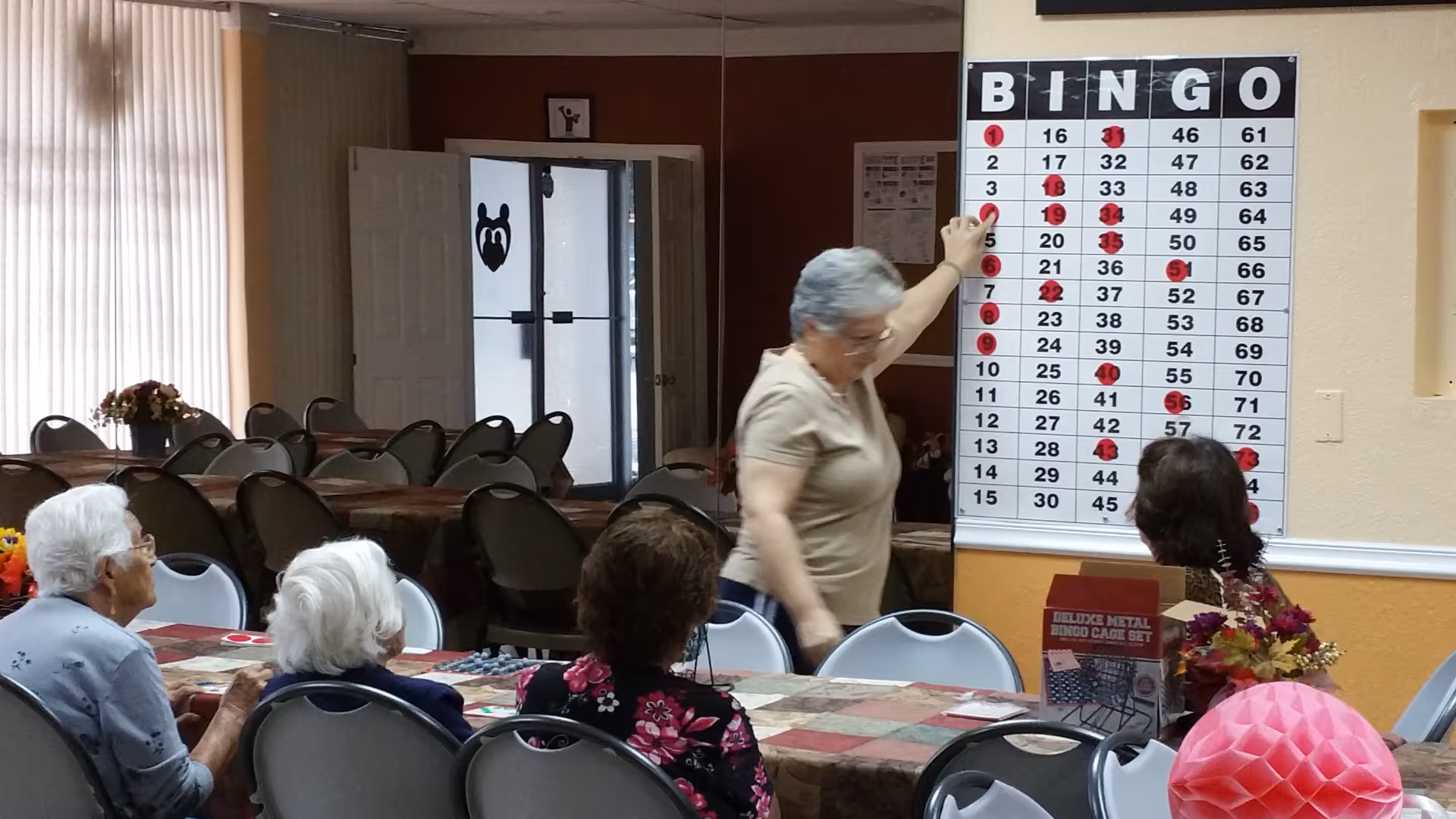 A group of elderly women seated around a table in a community room, playing bingo. One woman is standing and pointing to a large bingo board on the wall with marked numbers. The room has tables and chairs, with some floral decorations and a box labeled 'Deluxe Metal Bingo Cage Set' on the table.