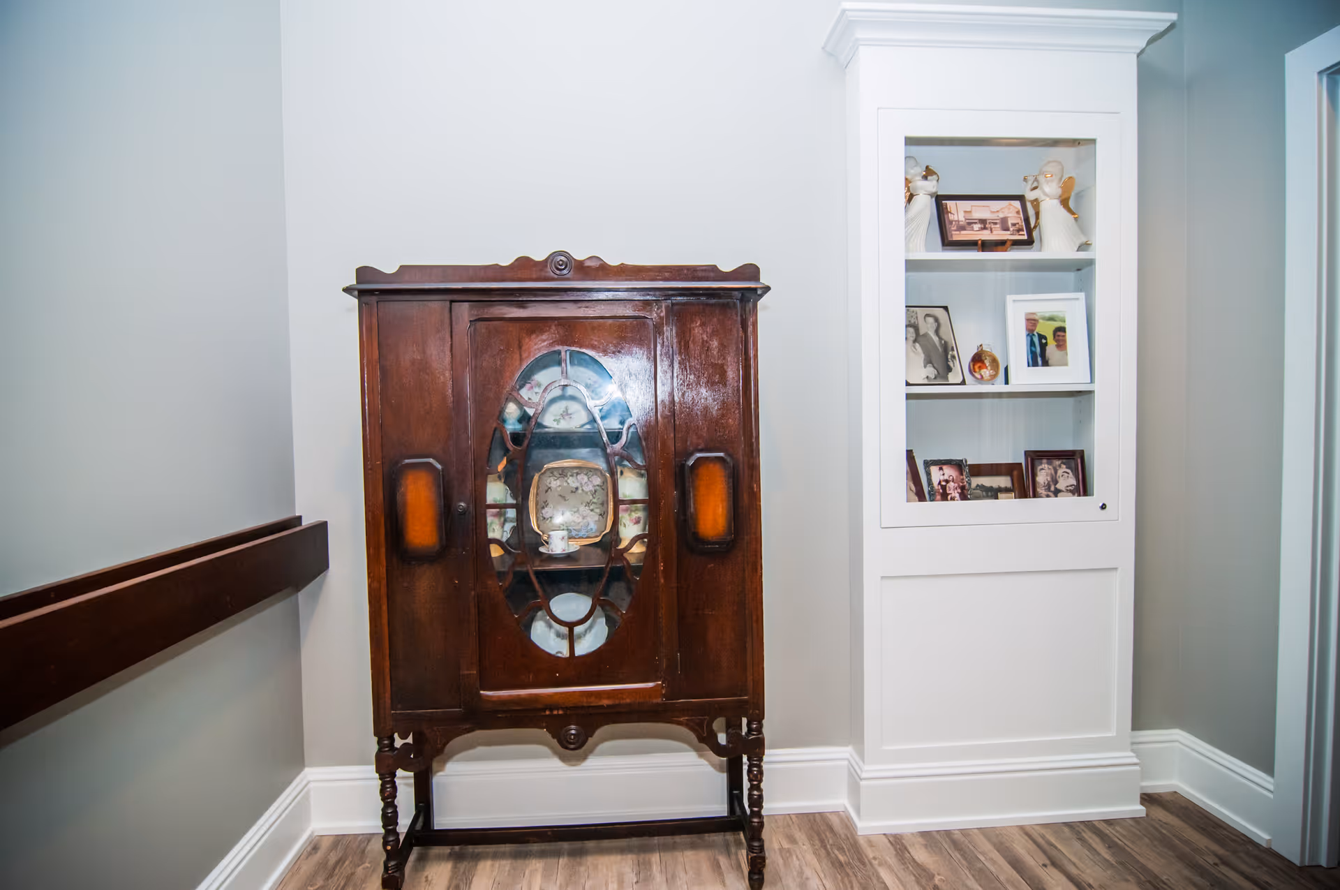 A corner of a room featuring a vintage wooden cabinet with glass doors displaying decorative plates, next to a white built-in shelving unit with framed photographs and angel figurines. The room has light gray walls and wood flooring.
