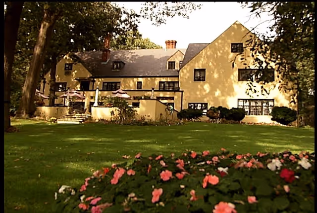 A large yellow two-story building with multiple windows and chimneys, surrounded by trees and a well-maintained lawn with pink and white flowers in the foreground.