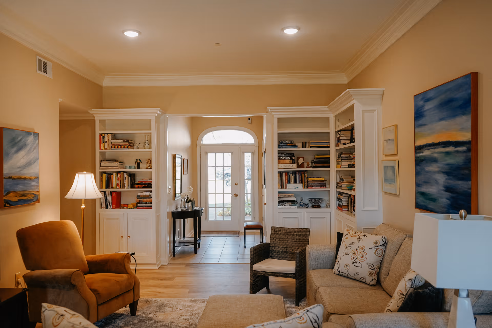 A cozy living room with beige walls and wooden flooring. The room features a comfortable beige sofa with patterned cushions, a brown armchair, a wicker chair, and a floor lamp. There are built-in white bookshelves filled with books on either side of a doorway with glass double doors leading outside. The walls are decorated with colorful landscape paintings.