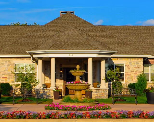 Front exterior view of a single-story building with a stone facade, a covered entrance supported by four columns, a central water fountain, benches on either side, and colorful flower beds in the foreground under a clear blue sky.