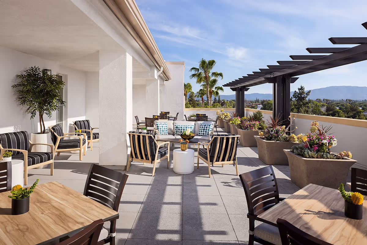 Outdoor patio area at Belmont Village Senior Living San Jose with multiple seating arrangements including cushioned chairs and sofas, wooden tables with chairs, large potted plants, and a pergola structure. The patio overlooks a scenic view with palm trees and mountains under a clear blue sky.