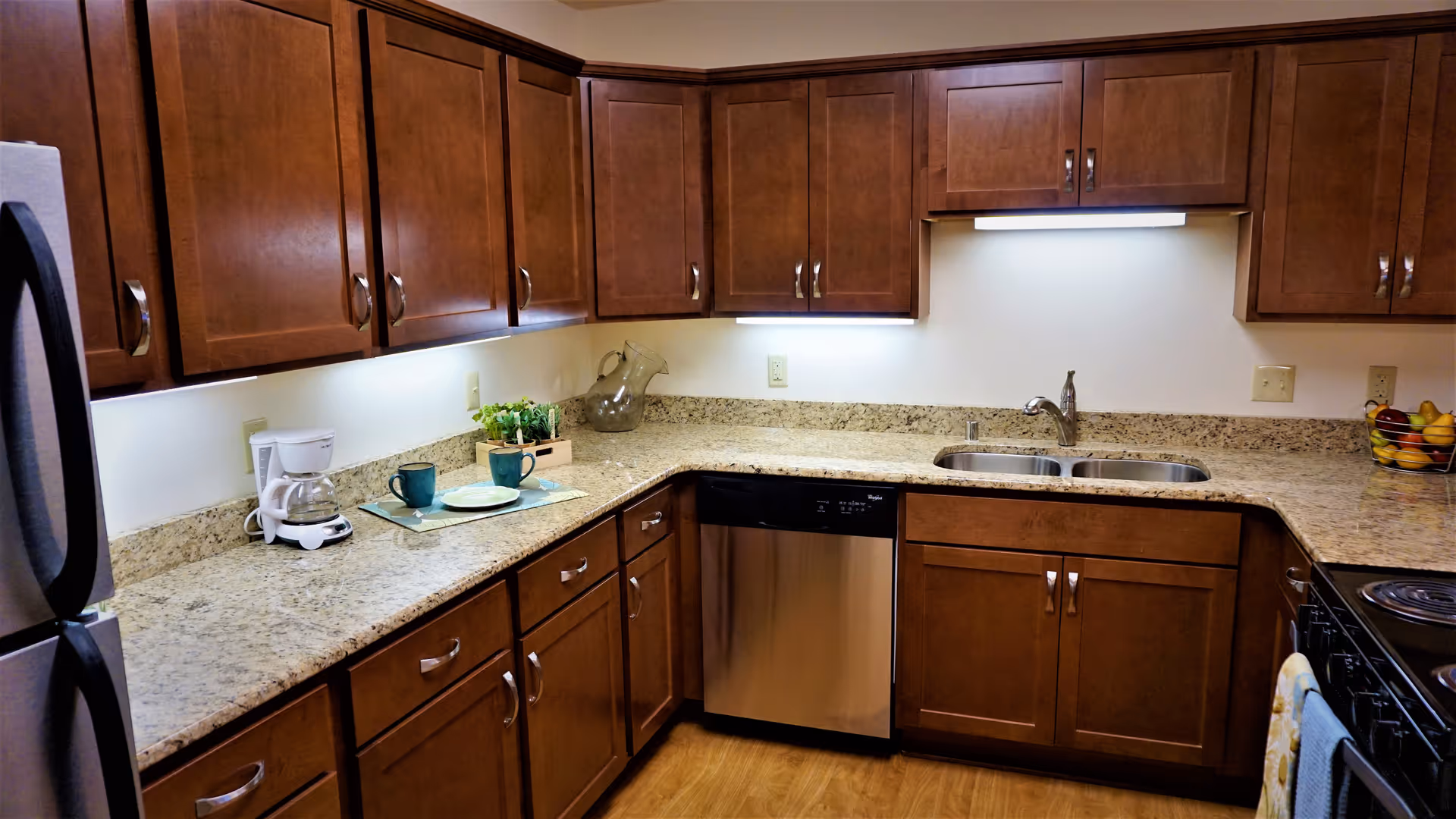 A modern kitchen with wooden cabinets, granite countertops, a stainless steel dishwasher, a double sink, a coffee maker, two blue mugs on placemats, a glass pitcher, and a fruit basket. The kitchen has under-cabinet lighting and a stove with a towel hanging on the handle.