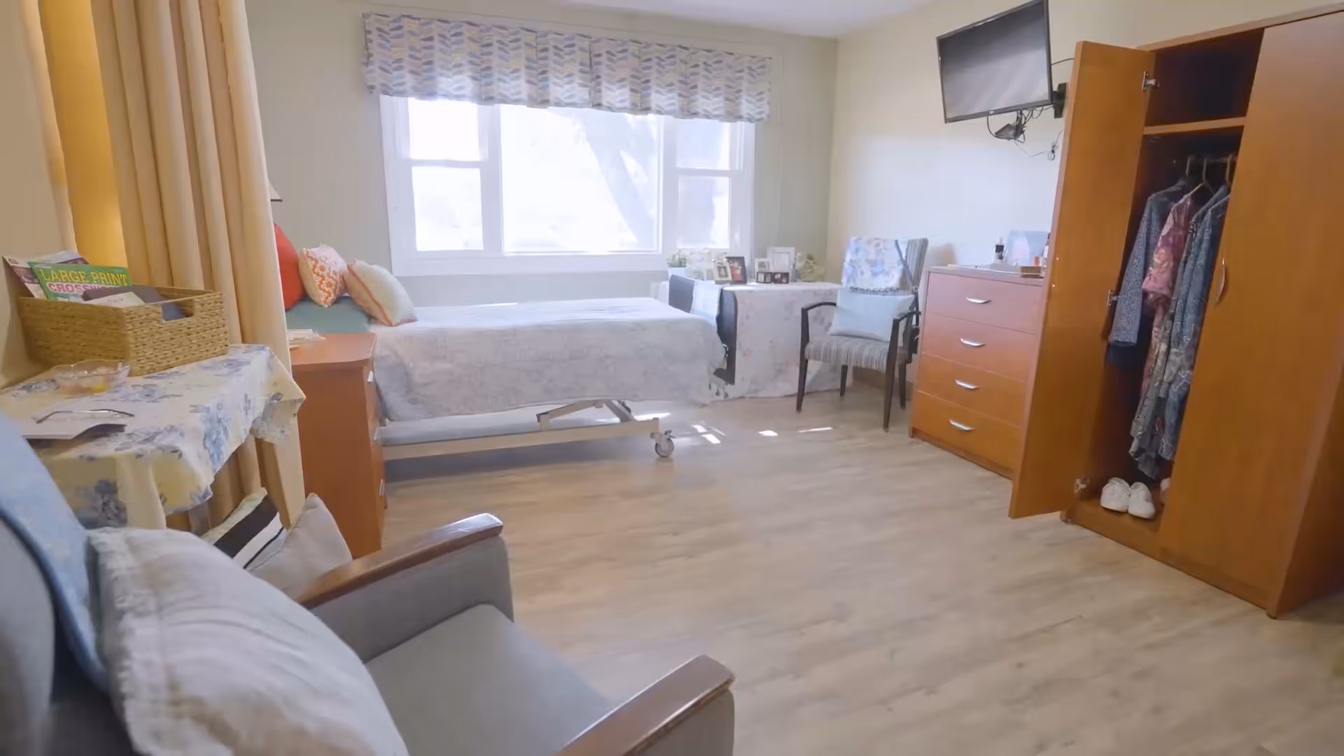 A bright and tidy bedroom in a senior living facility with a single bed near a window, a wooden wardrobe with clothes hanging inside, a chest of drawers, two chairs, a small table with a floral tablecloth, and a wall-mounted television.