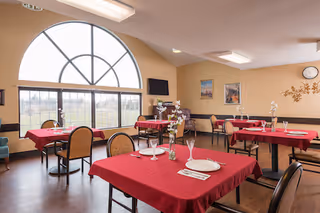 Dining room with several tables covered in red tablecloths, each set with white plates, glasses, and silverware. The room features a large arched window letting in natural light, beige walls with framed artwork, and a wall clock. Chairs with cushioned seats surround the tables.