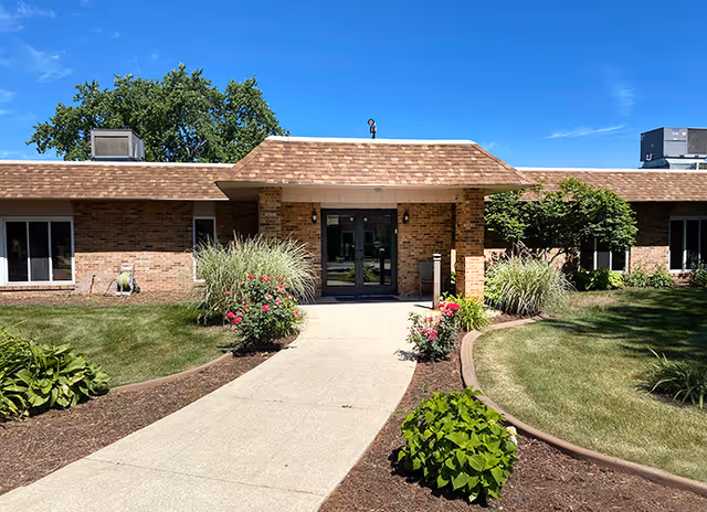 Front exterior view of Brickyard Healthcare - Portage Care Center building with a walkway leading to double glass doors, surrounded by green grass, bushes, and flowers under a clear blue sky.