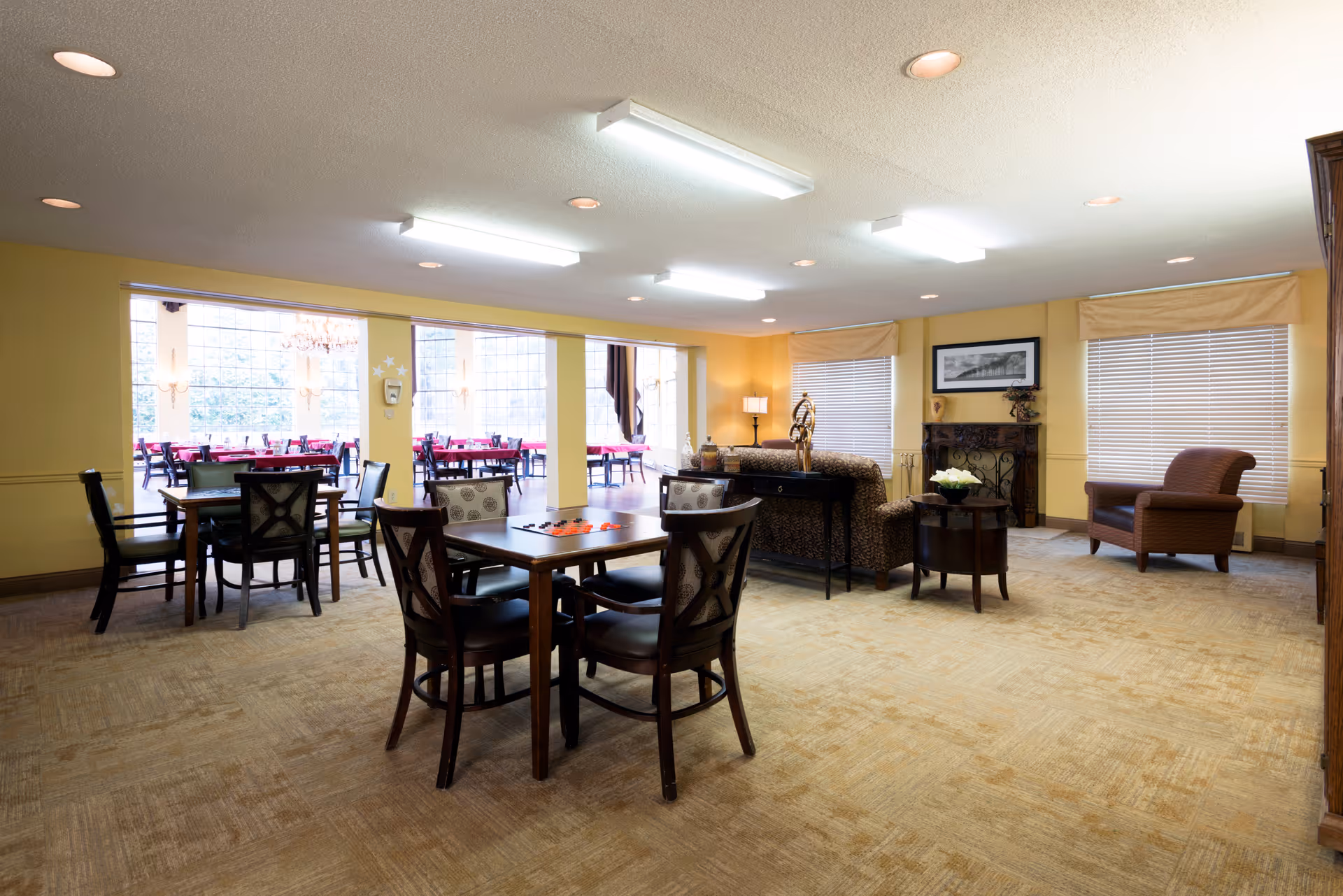 A spacious, well-lit common area in a senior living facility featuring several wooden tables with chairs arranged around them. The room has beige carpet flooring, yellow walls, and large windows with blinds. In the background, there is a seating area with a sofa, armchair, side tables, and decorative items including a framed picture and a lamp. Beyond the columns, another dining area with tables and chairs is visible through large windows.