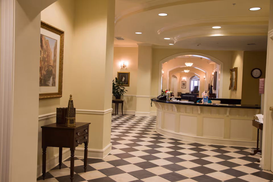 Interior view of a senior living facility reception area with a curved front desk, checkered black and white tile floor, beige walls, framed artwork, and decorative lighting fixtures.