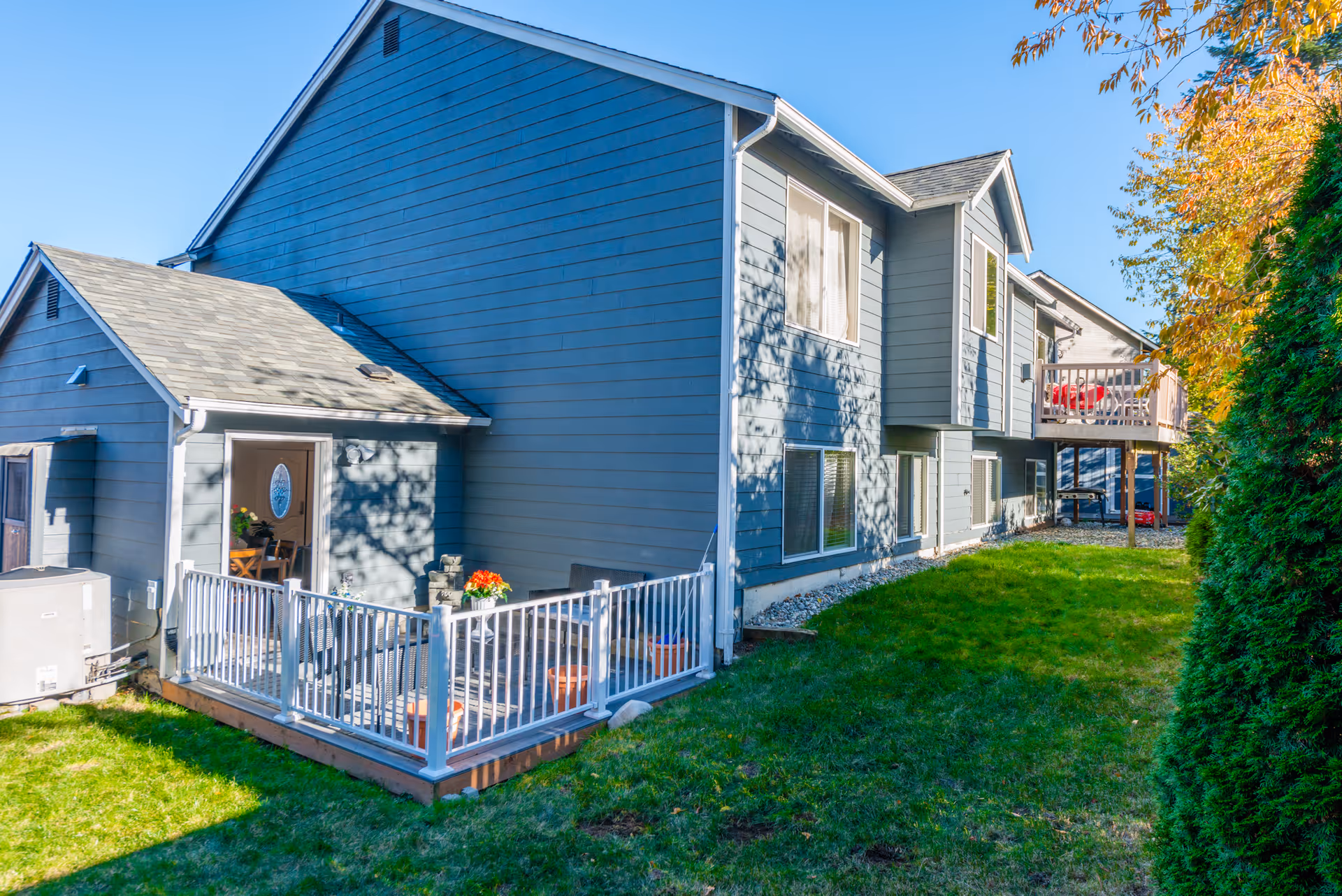 Exterior view of a two-story residential building with blue siding, a small fenced patio area with potted plants, and a grassy yard. The building has multiple windows and a small balcony on the upper floor. Trees with autumn-colored leaves are visible on the right side.