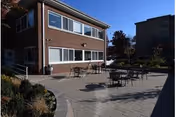 Outdoor patio area with several tables and chairs arranged on a paved surface next to a two-story brick building under a clear blue sky. There are some plants and trees around the patio, and another building is visible in the background.