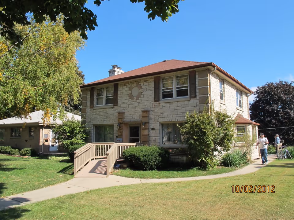 Exterior view of a two-story stone building with a brown roof and a wheelchair accessible ramp leading to the front door. The building is surrounded by green grass, bushes, and trees under a clear blue sky. Two people are walking on the sidewalk near the building.