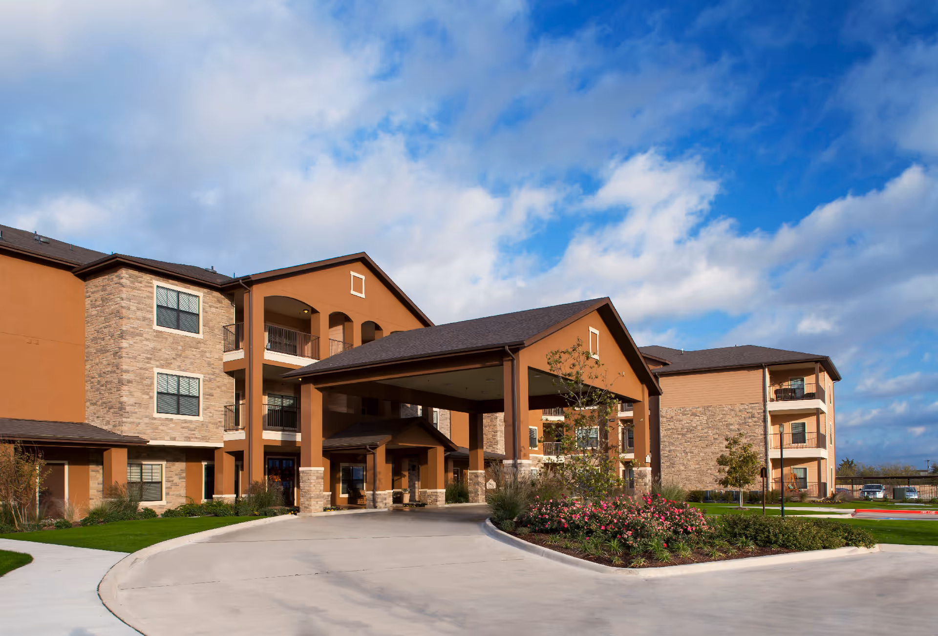 Exterior view of Sedona Place, a multi-story senior living facility with a covered entrance, stone and brown stucco facade, landscaped greenery, and a partly cloudy blue sky.