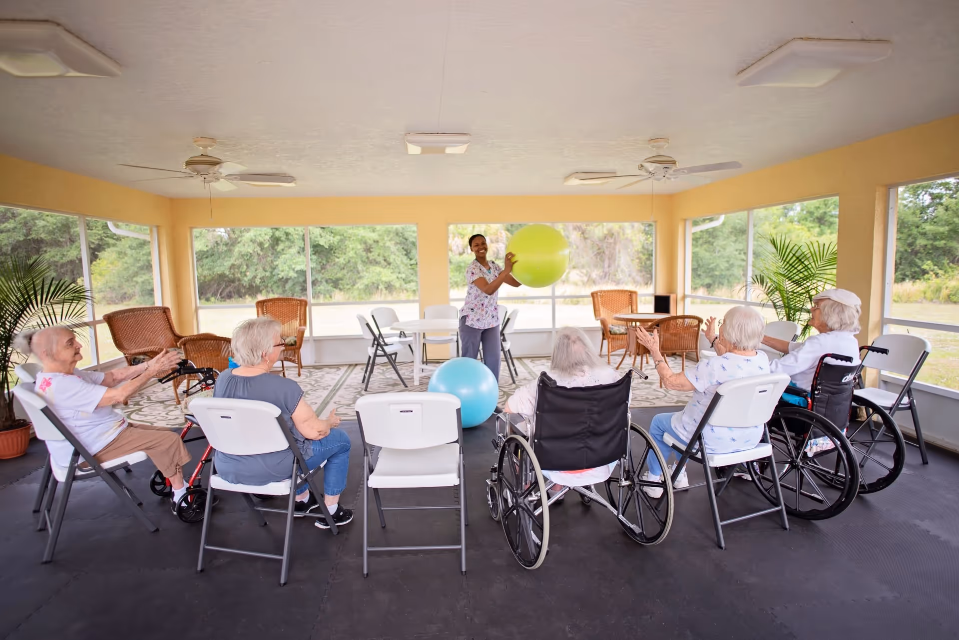 A group of elderly people seated in a semi-circle in a bright, screened-in room with large windows showing greenery outside. A caregiver stands in front of them holding a large yellow exercise ball, engaging the group in a recreational activity. Some participants are in wheelchairs, and others sit on folding chairs. The room has ceiling fans and wicker furniture in the background.