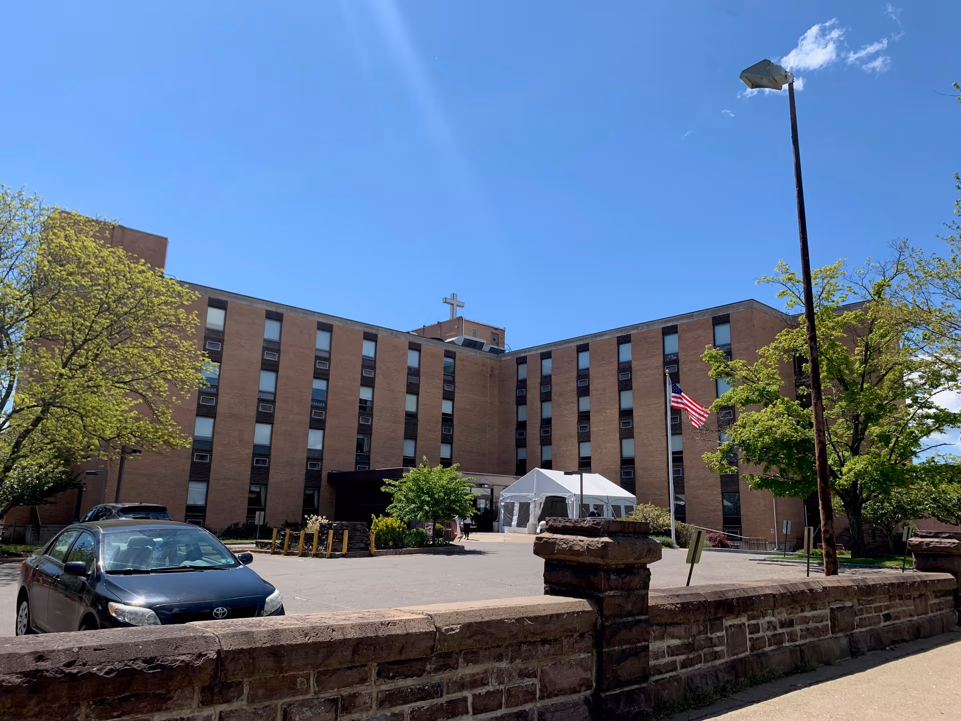 Exterior view of a multi-story brick building with numerous windows, an American flag on a flagpole, a white tent near the entrance, a parked black car, and trees with green leaves under a clear blue sky.