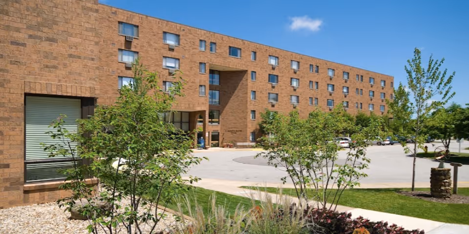Exterior view of a multi-story brick building with several windows, surrounded by green trees and shrubs under a clear blue sky. There is a paved driveway and parking area in front of the building.