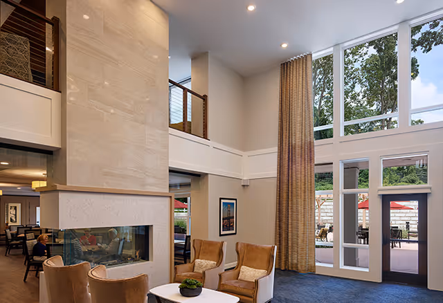 A bright and spacious senior living facility common area with high ceilings, large windows, and a modern double-sided fireplace. The room features comfortable armchairs arranged around a small table with a plant, and there is an outdoor patio visible through the glass doors and windows.