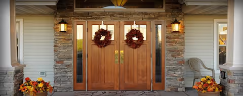 Entrance of a building with double wooden doors decorated with two autumn wreaths. The doors are flanked by stone walls and two wall-mounted lantern lights. There are flower pots with fall-themed flowers on either side of the entrance and a plastic chair to the right.