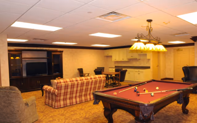 A cozy recreational room featuring a pool table with balls and cues set up, a plaid sofa facing a dark wooden entertainment center with a TV, several chairs around a small table, and a kitchenette area with cabinets and a countertop. The room is warmly lit with ceiling lights and a decorative hanging lamp above the pool table.