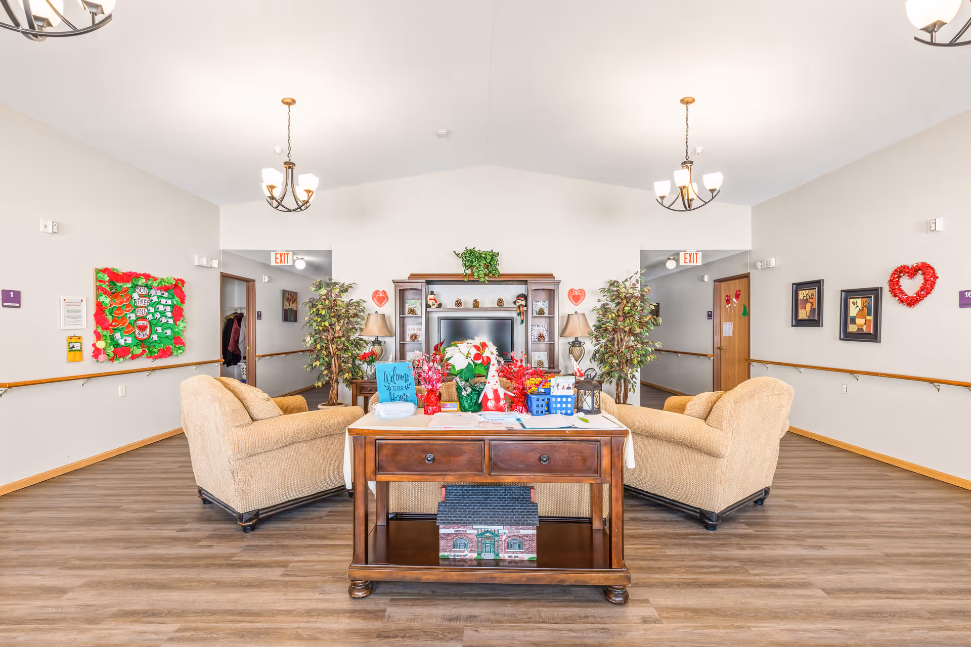 A bright and spacious living room area in a senior living facility with two beige armchairs facing a wooden entertainment center. The entertainment center has a TV, decorative items, and plants. A wooden table with drawers is placed between the chairs, holding various colorful decorations and papers. The room has light-colored walls with handrails, framed pictures, and heart-shaped decorations. Two chandeliers hang from the ceiling, and there are doorways leading to other rooms.