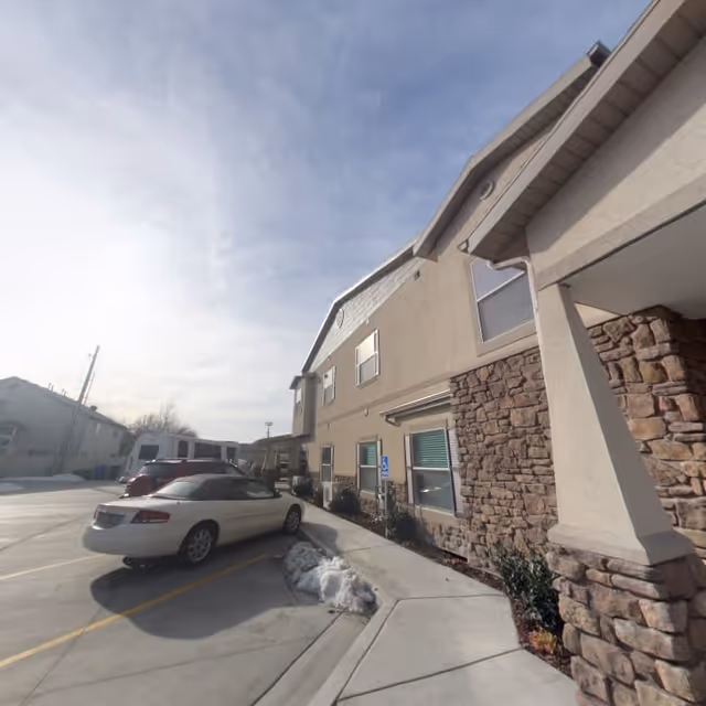 Exterior view of a two-story building with stone and beige siding, a sidewalk, and parked cars in a parking lot under a partly cloudy sky.