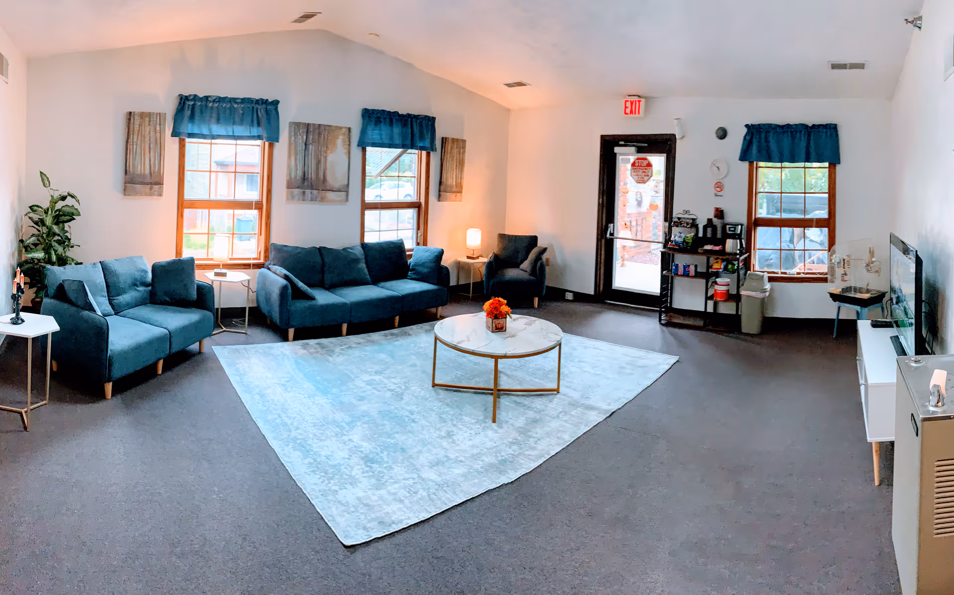 A bright and spacious living room with three blue upholstered sofas arranged around a light blue area rug and a round coffee table with a flower arrangement. The room has three windows with blue valances, a door with an exit sign above it, and various small tables and lamps. There is a TV mounted on the right wall and a small refreshment station near the door.