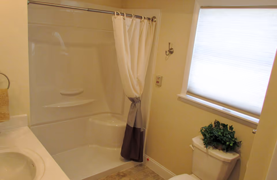 A clean bathroom with a white bathtub and shower combination featuring a white and gray shower curtain. There is a white toilet with a green plant on top, a window with a closed white blind, and a beige wall with a towel ring holding a beige towel. The floor has light-colored tiles.