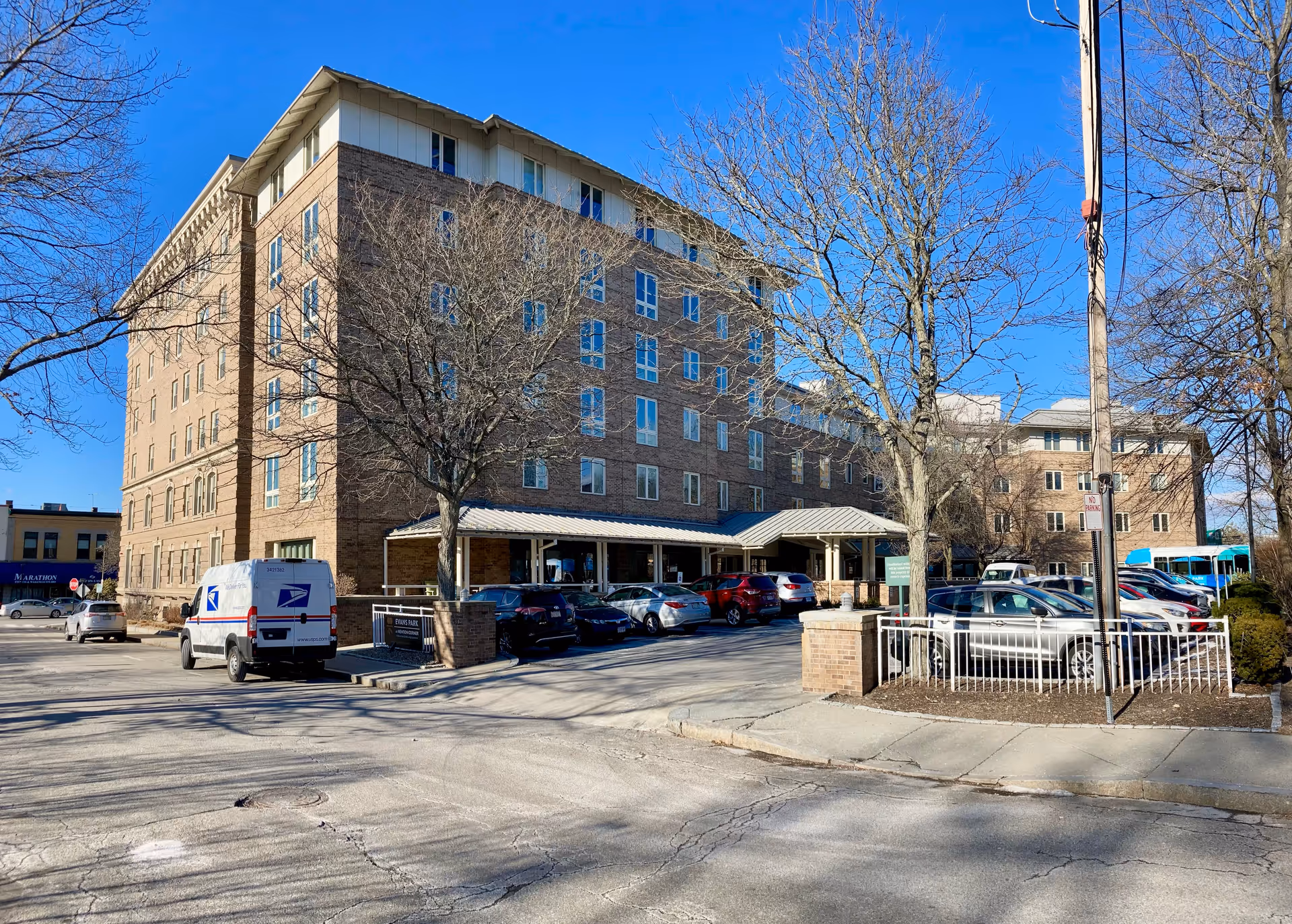 Exterior view of a multi-story brick building with several windows, a covered entrance, and a parking lot with cars and a USPS delivery van. Leafless trees are visible around the parking area under a clear blue sky.