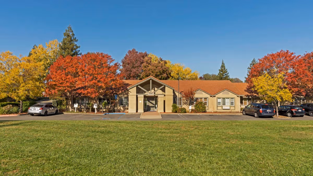 Front exterior view of a single-story building with a red-tiled roof surrounded by trees with autumn foliage in red, yellow, and orange. Several cars are parked in front of the building under a clear blue sky.