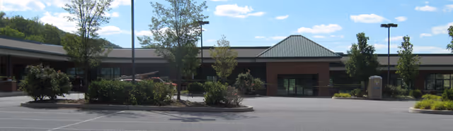 Exterior view of a single-story brick building with a green metal roof, surrounded by a parking lot with some trees and landscaped areas under a blue sky with scattered clouds.