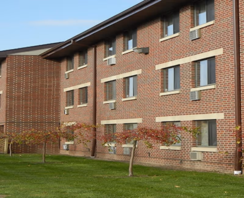 Exterior view of a three-story brick building with multiple windows and small trees with red leaves in front, set on a green lawn under a clear sky.