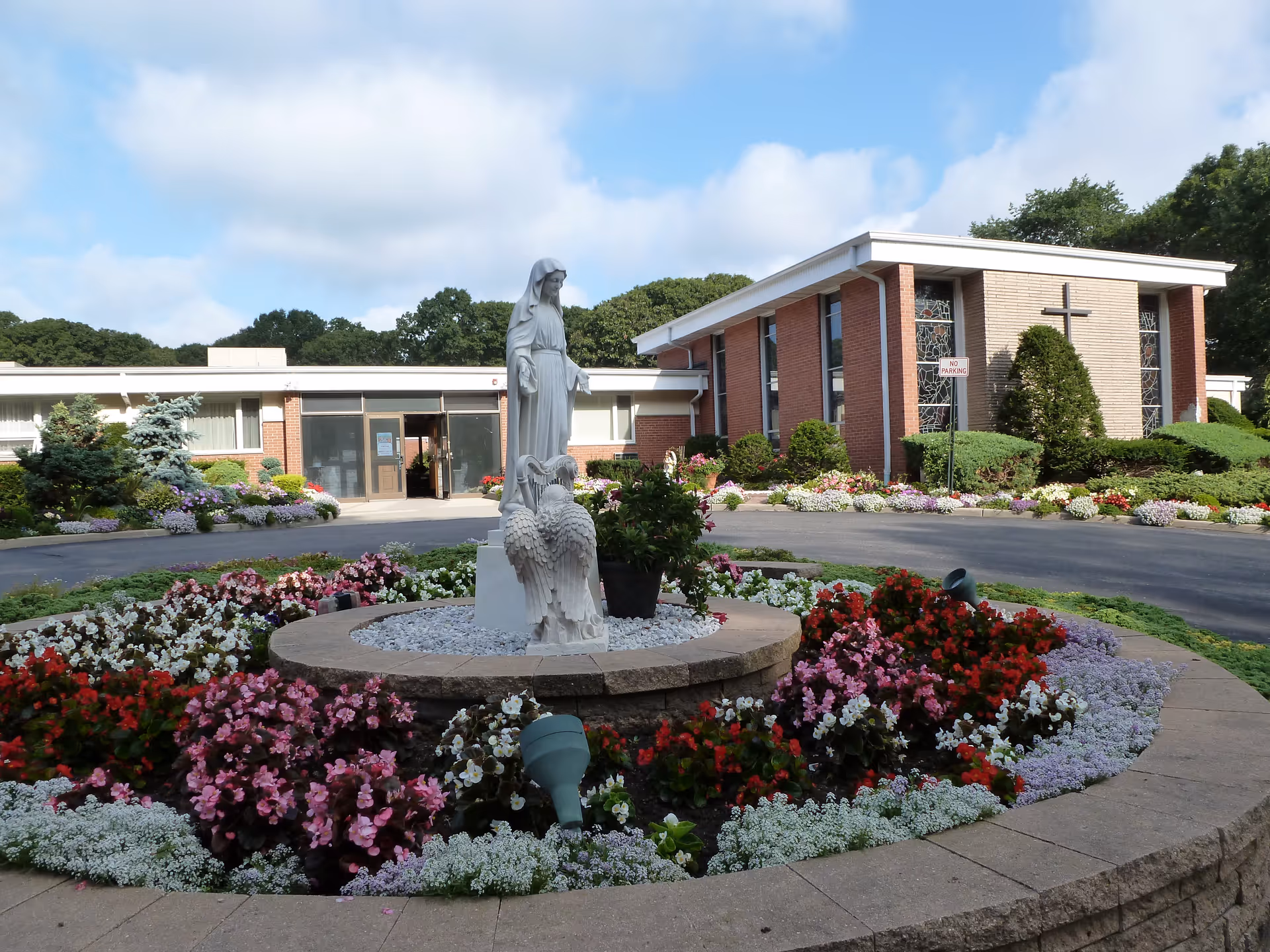 A circular flower bed with colorful flowers and a white statue of a religious figure in the center, surrounded by a paved driveway. In the background, there is a brick building with large windows, stained glass, and a cross mounted on the wall. The sky is partly cloudy.