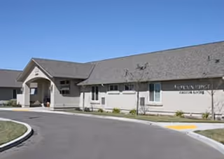 Exterior view of a single-story building with a gray roof and light-colored walls, featuring an entrance with a covered porch. The building has a sign that reads 'Autumn Ridge Assisted Living'. The driveway curves in front of the entrance, and there is a small landscaped area with grass and plants.