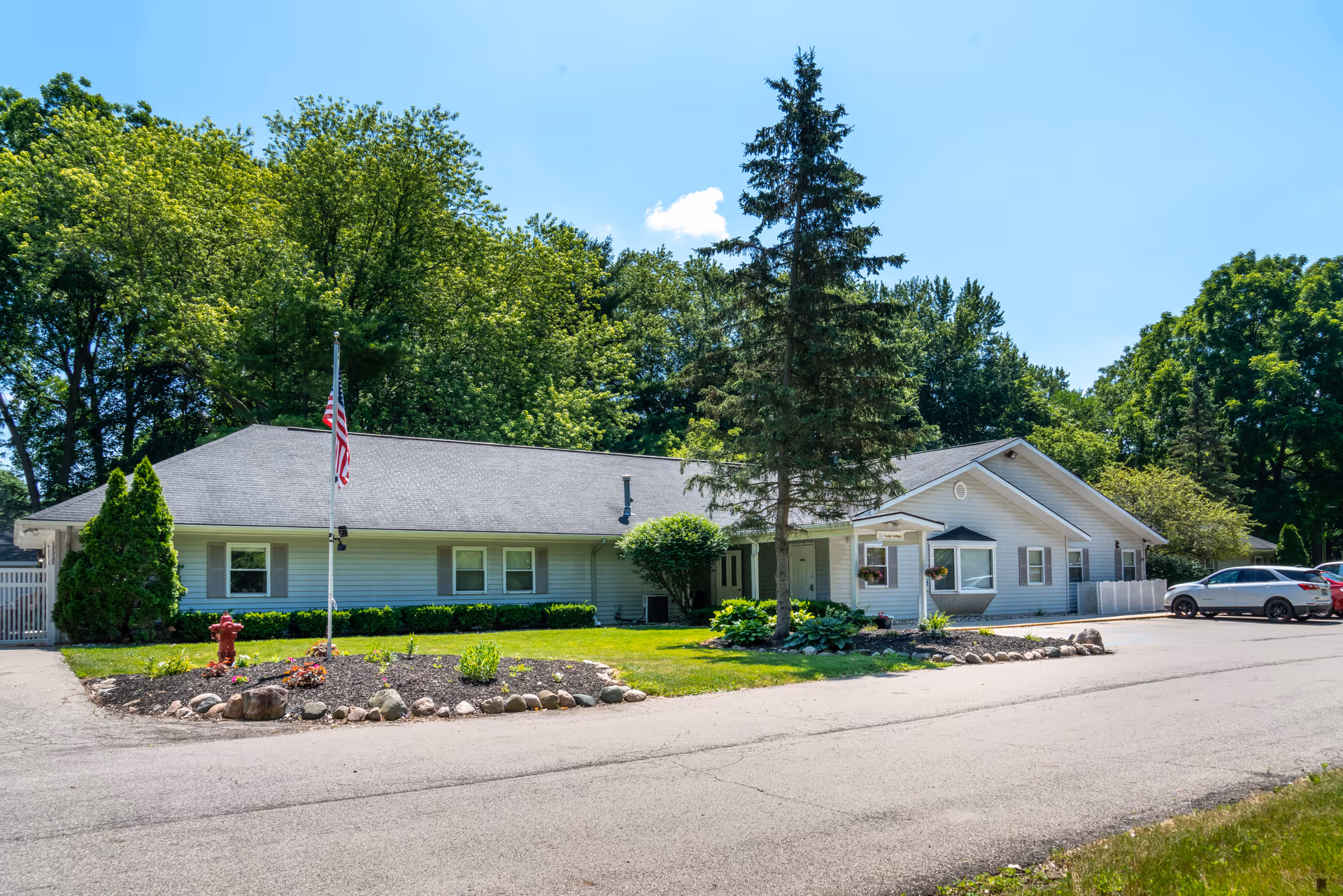 Single-story white building with a gray roof surrounded by green trees and a well-maintained lawn. There is a flagpole with an American flag, a fire hydrant, and several parked cars on the right side. The sky is clear and blue.