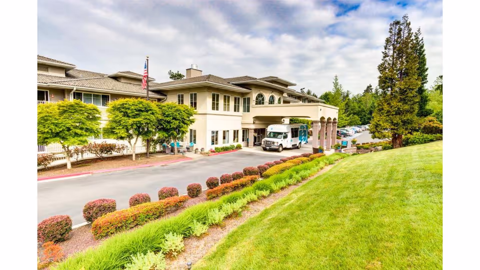 Exterior view of Harbour Pointe Senior Living facility showing a two-story building with a covered entrance, a parked shuttle bus, well-maintained landscaping with bushes and trees, and a green lawn under a partly cloudy sky.