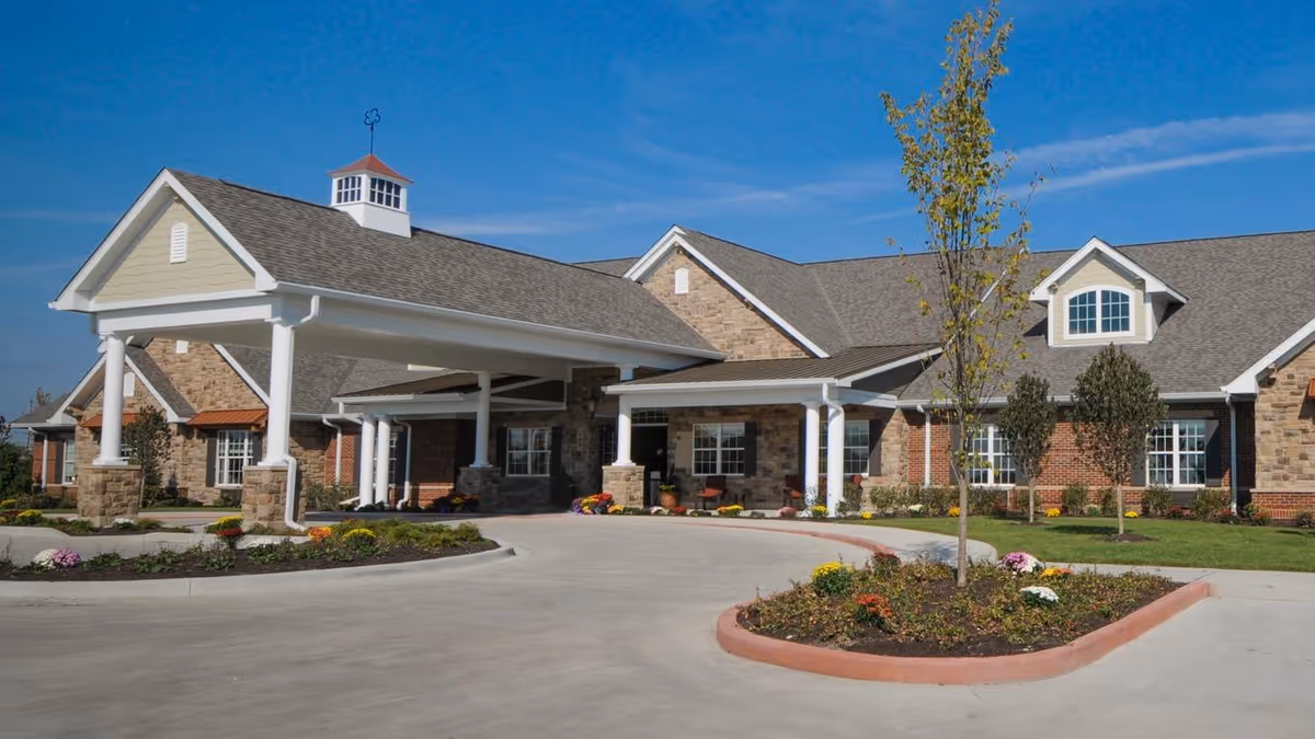 Exterior view of a senior living facility building with a covered entrance, stone and brick facade, landscaped flower beds, and a clear blue sky.