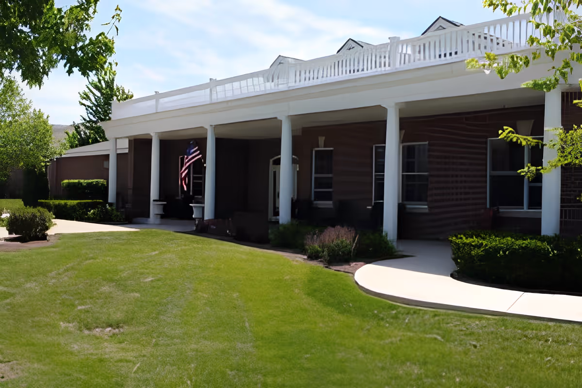 Exterior view of a single-story brick building with white columns and a white railing on the roof. There is a well-maintained green lawn and bushes in front of the building, with a concrete walkway leading to the entrance. An American flag is displayed near the entrance under the covered porch.
