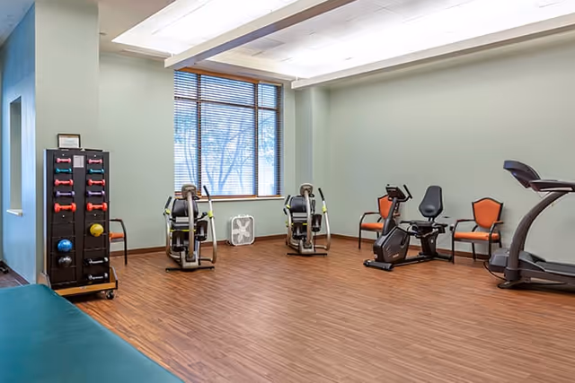 A small fitness room with wooden flooring and light green walls. The room contains exercise equipment including a treadmill, a recumbent bike, two arm exercise machines, a rack with colorful dumbbells and medicine balls, and two orange chairs. A window with blinds lets in natural light, and a white fan is placed on the floor in front of the window.