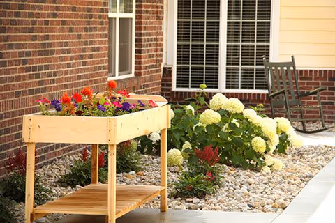 Outdoor garden area next to a brick building wall featuring a wooden planter box with colorful flowers, white hydrangea bushes, other small plants, a rock bed, and a dark green rocking chair near a window with white blinds.