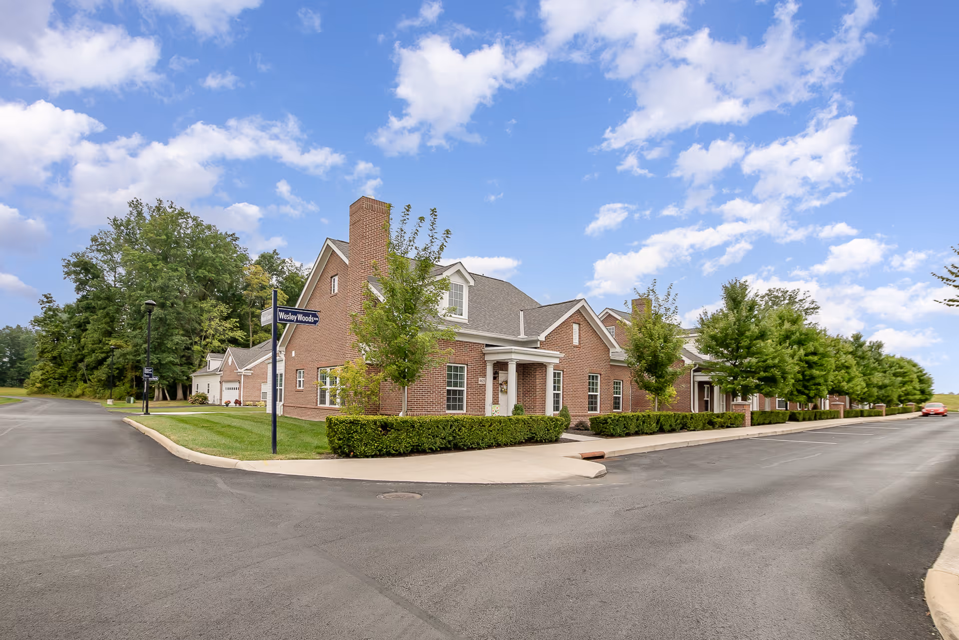 Exterior view of a brick building with a gray roof and multiple windows, surrounded by neatly trimmed bushes and trees along a paved road under a partly cloudy blue sky. A street sign reads 'Wesley Woods'.