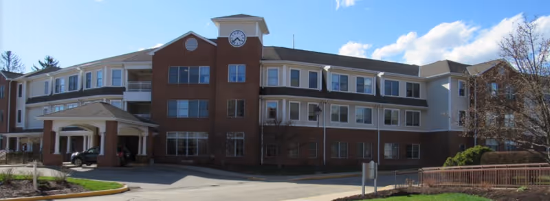 Exterior view of a three-story senior living facility building with a brick and beige facade, featuring a clock tower above the main entrance and several windows. The entrance has a covered drop-off area with a car parked underneath. The surrounding area includes a driveway, some landscaping with grass and trees, and a clear blue sky.
