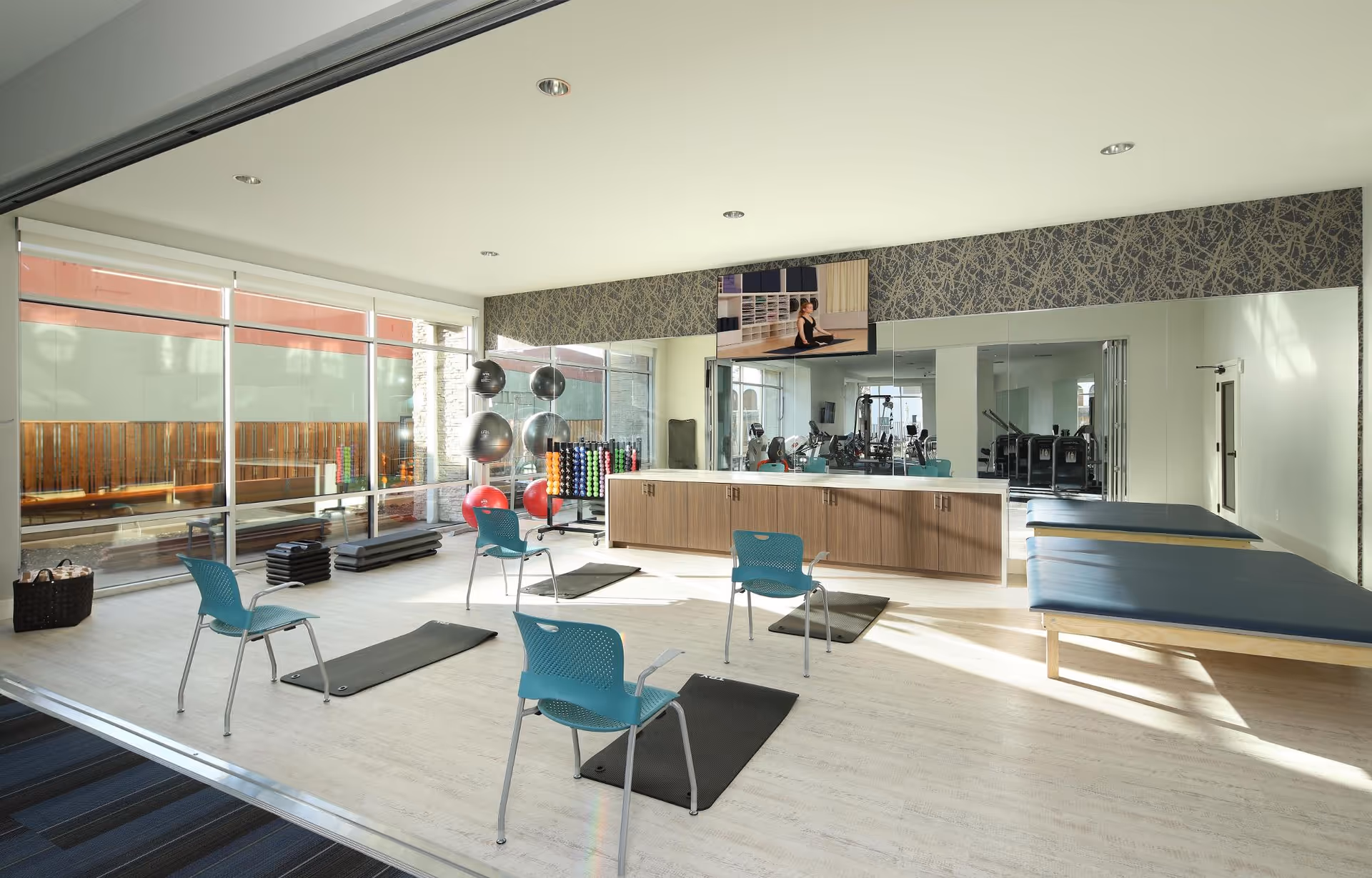 A bright and spacious exercise room with large windows letting in natural light. The room contains exercise mats on the floor, blue chairs, a rack of colorful dumbbells, exercise balls, and two padded treatment tables. A large mirror covers one wall, and a TV mounted above the cabinets shows a person doing yoga.